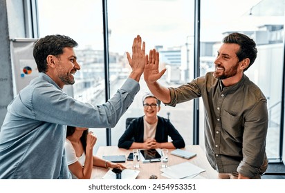 Profitable collaboration. Two middle age men standing by opposite sides of table and giving high five with smile. Joyful coworkers getting best results in department and making celebration gesture.