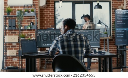 Programer coding on laptop sitting down at desk with computer screens parsing code in software agency. Software developer compiling algorithms with cloud programers working in the background.