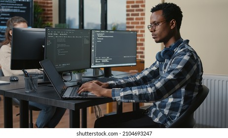 Programer thinking while touching beard and fixing glasses while typing on laptop sitting at desk with multiple screens parsing code. Focused database admin working with team coding in the background. - Shutterstock ID 2104693247