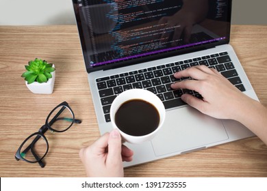 The programmer created some systems on the computer with the left hand holding a coffee cup on a brown wood desk. Top view.