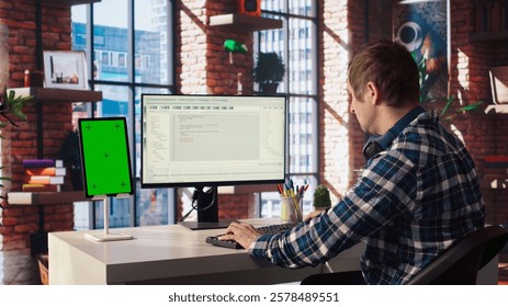 Programmer at desk typing code into computer, developing software programs, consulting green screen tablet. Man in home office fixing errors in application using chroma key device and PC, camera A