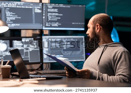 Programmer examines paperwork with serious expression and compares notes to code displayed on surrounding monitors. Developer with clipboard glancing at large displays of data, ensuring accuracy.