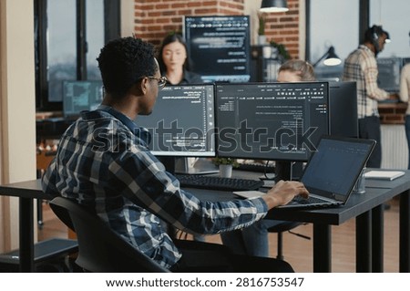 Programmer fixes his glasses while typing on a laptop and checking compiled code results at a desk with multiple software screens.