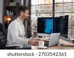 Programmer in glasses working on computer with dual monitors and laptop at table in office