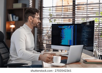 Programmer in glasses working on computer with dual monitors and laptop at table in office