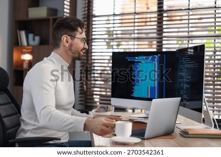Programmer in glasses working on computer with dual monitors and laptop at table in office