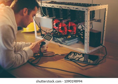 Programmer holding a wattmeter measuring cryptocurrency mining rig electrical energy consumption. Selective focus on the wattmeter - Shutterstock ID 1090198994