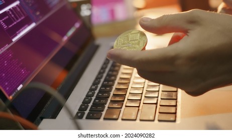 The programmer looks at the screen of the cryptocurrency mining graphics and turns bitcoin in his hands. A hacker's hand with bitcoin on the background of monitors with graphs of information at night.