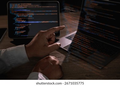 Programmer pointing at laptop screen with visible lines of code and tablet beside, concept of coding, software development, IT, cybersecurity, and digital technology innovation.