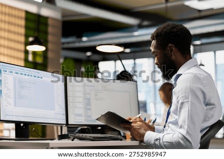 Programmer reviews documentation while coding on PC in modern office. Software developer cross referencing paperwork and source code at workstation to ensure accurate implementation