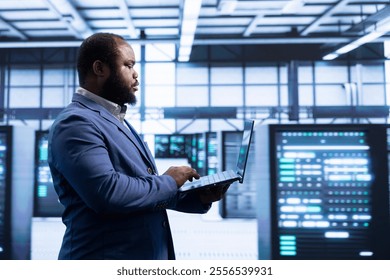 Programmer using laptop in data center with advanced technology, ensuring cybersecurity and efficient network management. Professional in server hub optimizing data center infrastructure with notebook