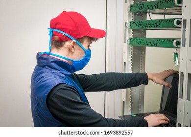 Programmer In Virus Protection Tools Works In The Server Room Datacenter. Maintenance Of Computer Equipment Under Quarantine.A Guy In Medical Mask Is Working In Front Of A Laptop.