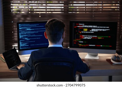 Programmer working on computer with dual monitors and laptop at table in office, back view