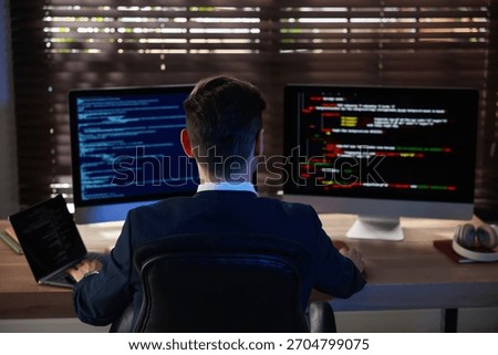 Programmer working on computer with dual monitors and laptop at table in office, back view