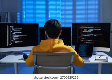 Programmer working on computer with triple monitors and laptop at white table in office, back view