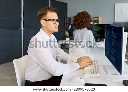 Programmers developers coding software teamwork in office. A focused engineer types at a computer with code on a large monitor in a tidy workplace. Strong team focus delivers better code.