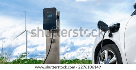 Progressive combination of wind turbine and EV car, future energy infrastructure. Electric vehicle being charged at charging station powered by renewable energy from wind turbine in the countryside.