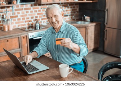 Progressive innovations. Joyful nice elderly man holding a credit card and making an online payment while using his laptop