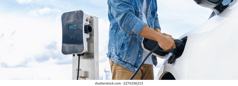 Progressive man with his electric car, EV car recharging energy from charging station on green field with wind turbine as concept of future sustainable energy. Electric vehicle with energy generator. - Shutterstock ID 2276106213