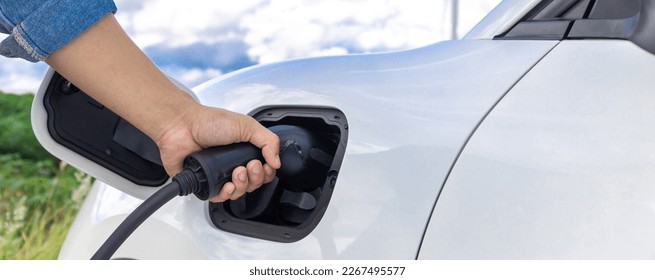 Progressive natural scenic with windmill generator where hand insert charging plug to electric vehicle from charging station with natural background. EV car powered by wind turbine electric generator. - Shutterstock ID 2267495577