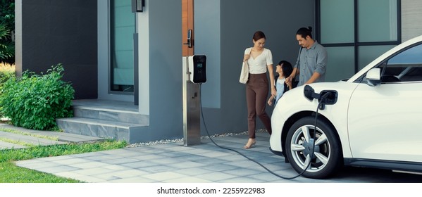 Progressive young parents and daughter with electric vehicle and home charging station. Green and clean energy from electric vehicles for healthy environment. Eco power from renewable source at home. - Shutterstock ID 2255922983