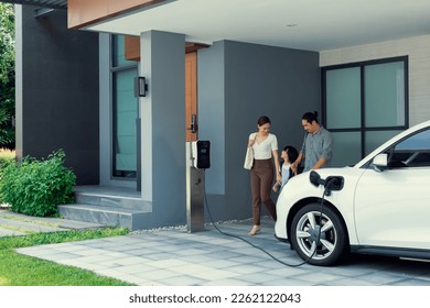 Progressive young parents and daughter with electric vehicle and home charging station. Green and clean energy from electric vehicles for healthy environment. Eco power from renewable source at home. - Shutterstock ID 2262122043