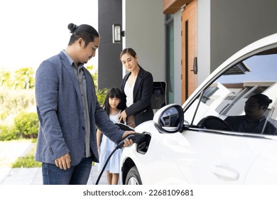 Progressive young parents and daughter with electric vehicle and home charging station. Green and clean energy from electric vehicles for healthy environment. Eco power from renewable source at home. - Shutterstock ID 2268109681