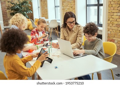 Project. Kind Young Female Teacher In Glasses Showing Scientific Robotics Video To Little Boy, Using Laptop In Classroom