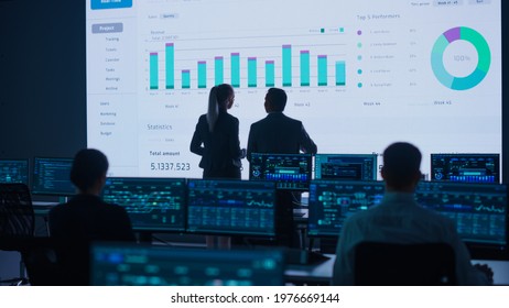 Project Leader, Chief Executive Discuss Data Shown on Big Display. Screens Show Infographics, Charts, Finance Analysis, Stock Market, Growth.Telecommunications Control Room with Working Professionals
