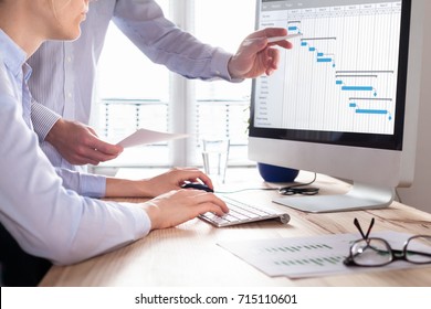 Project management team updating Gantt chart schedule or planning on computer, two business people in office