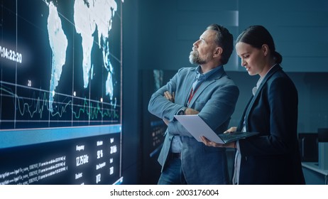 Project Manager and Computer Science Engineer Talk while Using Big Screen Display and a Laptop, Showing Infrastructure Infographics Data. Telecommunications Company System Control and Monitoring Room.