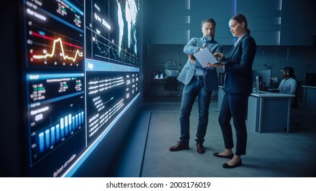 Project Manager and Computer Science Engineer Talk while Using Big Screen Display and a Laptop, Showing Infrastructure Infographics Data. Telecommunications Company System Control and Monitoring Room. - Shutterstock ID 2003176019