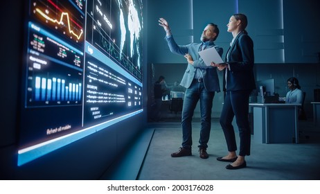 Project Manager and Computer Science Engineer Talk while Using Big Screen Display and a Laptop, Showing Infrastructure Infographics Data. Telecommunications Company System Control and Monitoring Room.
