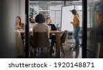 Project Manager Makes a Presentation for a Young Diverse Creative Team in Meeting Room in an Agency. Colleagues Sit Behind Conference Table and Discuss Business Development, User Interface and Design.