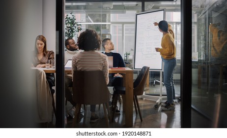 Project Manager Makes a Presentation for a Young Diverse Creative Team in Meeting Room in an Agency. Colleagues Sit Behind Conference Table and Discuss Business Development, User Interface and Design.
