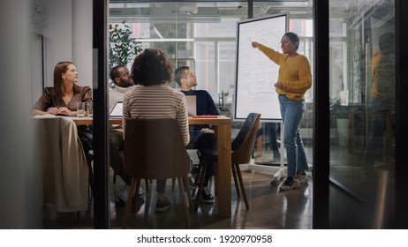 Project Manager Makes A Presentation For A Young Diverse Creative Team In Meeting Room In An Agency. Colleagues Sit Behind Conference Table And Discuss Business Development, User Interface And Design.