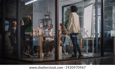 Project Manager Makes a Presentation for a Young Diverse Creative Team in Meeting Room in an Agency. Colleagues Sit Behind Conference Table and Discuss Business Development, User Interface and Design.