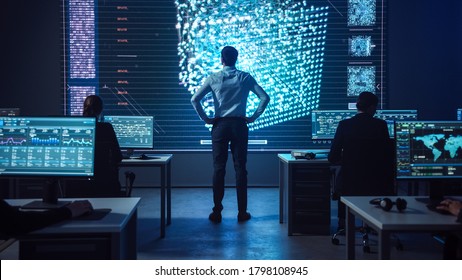 Project Manager Stands Next To Big Screen With Neural Network. Team Of Professional Computer Data Science Engineers Work On Desktops In A Dark System Control And Monitoring Telecommunications Office.