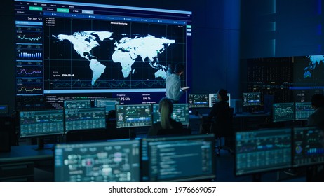 Project Manager Uses Digital Tablet Looks at Big Screen Display Showing Infographics and Global World Map Data. Telecommunications Control Monitoring Room with People Working