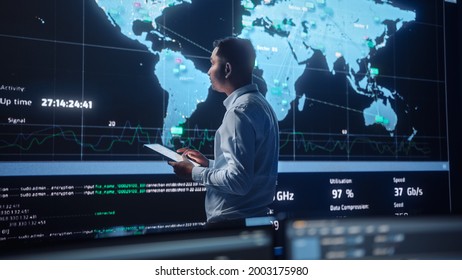 Project Manager Walks To A Big Screen With Global Map. He Is Using A Tablet. Team Of Professional Computer Data Science Engineers Work In A Dark System Control And Monitoring Telecommunications Office