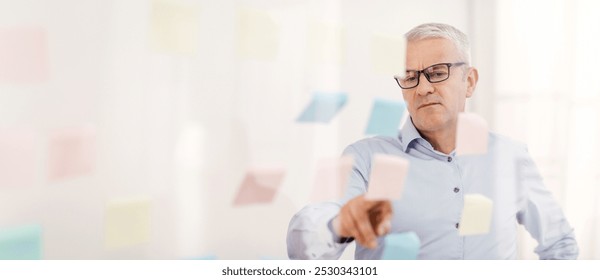 Project Planning. Serious Mature Businessman Reading Colorful Sticky Notes On Glass Visualisation Board In Office - Powered by Shutterstock