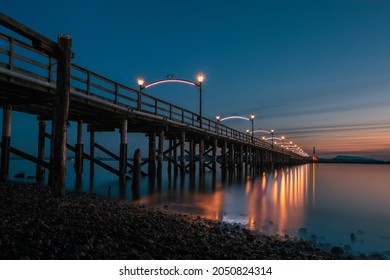 Promanade bridge Wooden pier at White Rock, BC, Canada extends diagonally into image. It's twinkling lights reflected in the sea. Sunset turns sky to blue and orange over the Gulf and islands