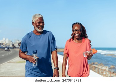 Promenade walk: An older black couple happily walking together along a seaside promenade, laughing and holding water bottles, illustrating active senior lifestyle and joy. - Powered by Shutterstock