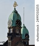 Prominent clock tower topped with a golden weather vane, featuring two large clocks with roman numerals set within a verdant patina dome.