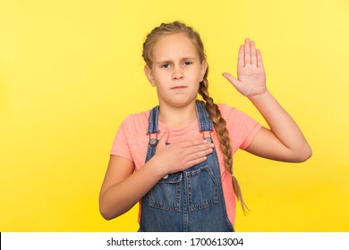 I Promise To Be Honest! Portrait Of Serious Responsible Little Girl With Braid In Denim Overalls Raising Palm To Take Oath, Child Swearing To Tell Truth. Studio Shot Isolated On Yellow Background