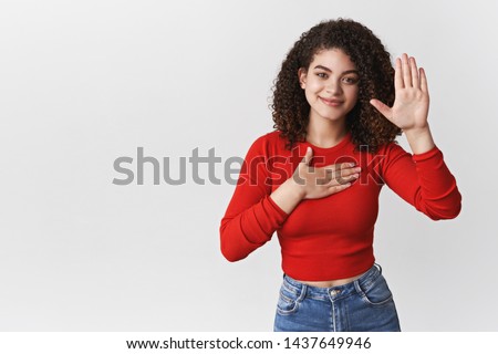 Promise tell truth. Charming happy relaxed sincere young curly-haired girl pleadging oath swear hold hand heart raise palm making statement smiling happily, standing white background