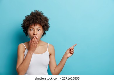 Promotion concept. Surprised curly haired woman covers mouth with hand points aside at copy space offers huge sale shows banner design dressed in casual white t shirt isolated over blue background