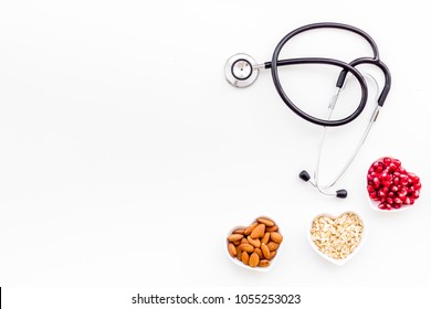 Proper nutrition for pathients with heart disease. Cholesterol reduce diet. Oatmeal, pomegranate, almond in heart shaped bowl near stethoscope on white background top view copy space
