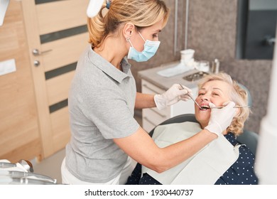 Proper Work. High Angle View Of The Senior Blonde-haired Woman Having Her Teeth Examined By Serious Doctor Wearing Protective Mask. Examination And Inspection At The Dental Clinic Concept. Stock Photo