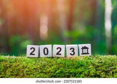 Property 2023 Concept. Wooden Cubes With Numbers 2023 And House Icon On Mossy Fallen Tree In Summer Forest In Focus. Background Blurred.